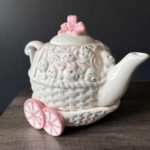 May include: A white ceramic teapot with a basket weave design, featuring smiling bunny faces and pink accents. The lid is topped with a pink bow, and the teapot rests on a base with two pink wheels. A charming Easter-themed collectible.