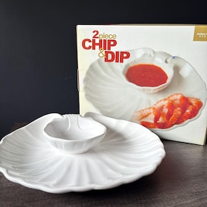 May include: A white ceramic two-piece chip and dip serving set shaped like a large scallop shell with a smaller integrated bowl. The set is displayed in front of its original cardboard packaging featuring the text 2 piece CHIP & DIP.
