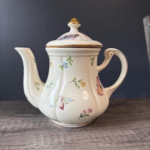 May include: A cream-coloured porcelain teapot with a fluted body and a matching lid. The teapot features a delicate pattern of colourful butterflies, bees, and small floral sprigs. The handle, spout, and lid finial are accented with gold trim.