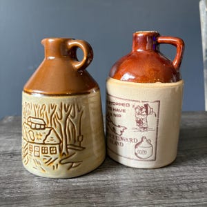 Peut inclure: Deux pichets en céramique avec des hauts et des anses émaillés marron. Le pichet de gauche présente un relief moulé représentant une cabane dans une forêt. Celui de droite a une base crème avec un motif marron imprimé d'une personne, d'une carte et du texte "STOPPED TO HAVE A NIP" et "PRINCE EDWARD ISLAND".