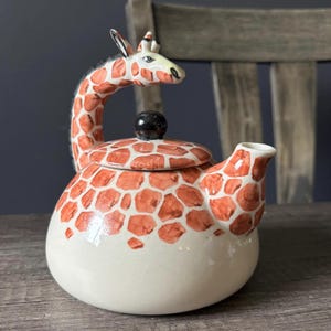 May include: A ceramic teapot featuring a giraffe design. The teapot is white with orange and brown giraffe spots. The handle is the giraffe's neck and head. The lid has a black knob.