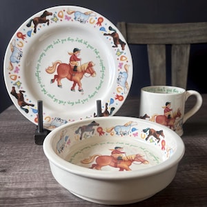 May include: A cream-coloured ceramic tableware set with a horse riding theme, including a plate, bowl, and mug. The plate and bowl feature a central image of a child riding a horse, surrounded by a border of horses and ribbons. The mug has the same image.