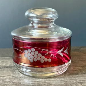 May include: A round glass jar with a matching lid, featuring a deep red translucent band around the centre. The red section is decorated with an etched white design of a grape cluster and vine leaves, providing a clear view of the glass.