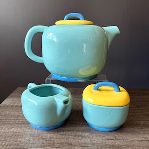 May include: A ceramic tea set consisting of a large teapot with a yellow lid and blue handle, a matching creamer, and a sugar bowl with a yellow lid. Each piece features a glossy light blue body with a blue base and rounded, smooth edges.