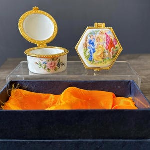 May include: Two decorative porcelain trinket boxes. One is round with a floral design and the other is hexagonal with a painted scene. Both have gold-coloured rims and hinges. The boxes are displayed on a clear stand, with an orange fabric in a blue box below.