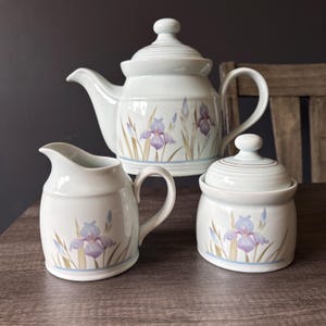 May include: A ceramic tea set consisting of a teapot with a lid, a creamer, and a sugar bowl with a lid. Each piece is off-white with a painted pattern of purple iris flowers and green stems, featuring a light blue band near the base.