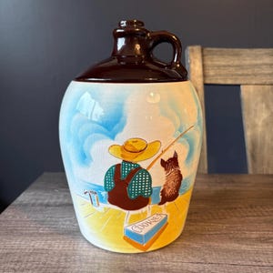May include: A vintage ceramic cookie jar shaped like a jug, with a brown lid and handle. The jar is decorated with a scene of a person fishing with a dog, and a cookie box. The jar is on a wooden surface.