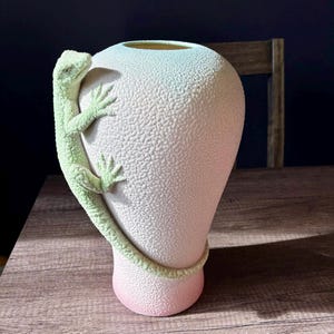 May include: A decorative vase with a textured surface and a gradient colour scheme, transitioning from pink at the base to white and light blue at the top. A light green lizard sculpture wraps around the vase, adding a unique artistic element.