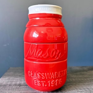 Op de afbeelding: Een stapelbare rode keramische maatbekerset in de vorm van een weckpot. De set bestaat uit drie delen met het woord "Mason" in reliëf op het middelste deel. Het onderste deel leest "Glassware Est. 1986". De deksel is wit.