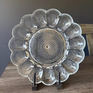 May include: Clear glass devilled egg serving platter with a scalloped edge and individual egg compartments. The centre of the platter has a textured, dotted design. The platter is displayed on a black metal stand.