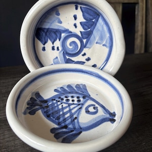 May include: Two round ceramic bowls with a white base and blue hand-painted designs. The front bowl features a stylised fish with cross-hatched details, while the back bowl displays a central spiral surrounded by leaf-like patterns and dots.