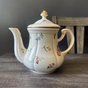 Peut inclure: Une théière en céramique blanche avec une poignée et des accents dorés. La théière est décorée d'illustrations colorées d'insectes, de fleurs et de feuillages. Le couvercle est surmonté d'un bouton doré.