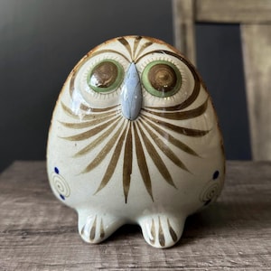 May include: A ceramic owl figurine with a cream-coloured base, featuring brown painted feather patterns radiating from a light blue beak. The owl has large green and brown eyes and small blue spiral accents on its sides, with two feet at the base.