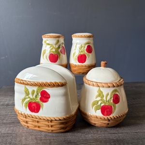 May include: A ceramic kitchen set featuring a napkin holder, a lidded sugar bowl, and two salt and pepper shakers. Each piece is cream-coloured with a painted red apple and green leaf motif, accented by a brown woven basket-style base and rim.