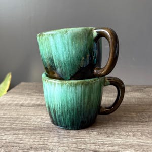 Op de afbeelding: Twee gestapelde keramische mokken met een glanzende, getextureerde afwerking. De mokken zijn diep zeegroen met bruine handvatten en accenten. Het glazuur heeft een druppeleffect, waardoor een uniek patroon ontstaat. De mokken staan op een houten oppervlak.