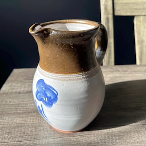 May include: A handmade ceramic pitcher with a brown rim and handle, and a white body. The pitcher features a blue floral design. The pitcher is sitting on a wooden surface.