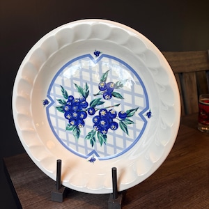 May include: A round white ceramic pie dish with a fluted edge. The centre features a blue lattice pattern overlaid with a detailed illustration of clusters of dark blue blueberries and green leaves. The design is centred on the white base.