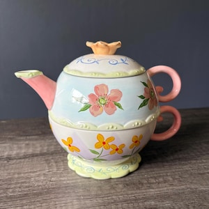 May include: A ceramic tea for one set featuring a teapot stacked on a teacup. The teapot has a light blue background with pink flowers and a pink spout. The teacup features a light purple diamond pattern with yellow flowers and a pink handle.
