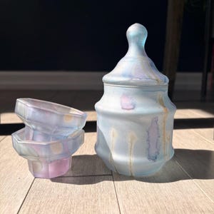 May include: A frosted glass jar with a matching lid and two small matching bowls. The set features a soft, translucent finish in shades of pale blue, lavender, and cream, with a subtle marbled or watercolour effect across the smooth glass surfaces.
