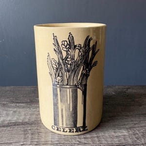 May include: A cylindrical ceramic utensil holder in a light beige colour featuring a black graphic print of a bunch of celery stalks inside a container. The word CELERY is printed in black capital letters at the base of the illustration.