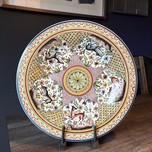 May include: A round ceramic decorative plate featuring a central green floral medallion surrounded by five scalloped panels. Each panel depicts a deer or stag in a natural setting with flowers and foliage, set against a yellow lattice background.