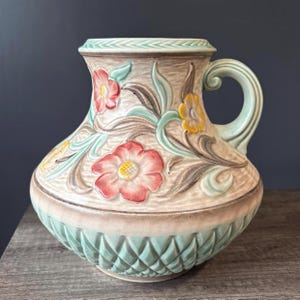 Może przedstawiać: Ceramiczny dzbanek o teksturowanym beżowym korpusie z tłoczonymi czerwonymi i żółtymi kwiatami, zielonymi liśćmi oraz jasnym zielonym uchwytem. Dolna część posiada podstawę w romby, a krawędź ozdobny wzór w formie warkocza.