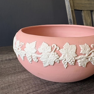 May include: A round, pink ceramic bowl with a white relief design of grape vines and leaves. The bowl sits on a wooden surface. The background is dark, with a wooden chair visible.