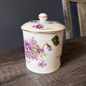 May include: A white porcelain lidded jar featuring a delicate pattern of purple violet flowers and green leaves. The cylindrical container has a matching lid with a small knob handle, and both the rim and lid edge are accented with a thin gold trim.