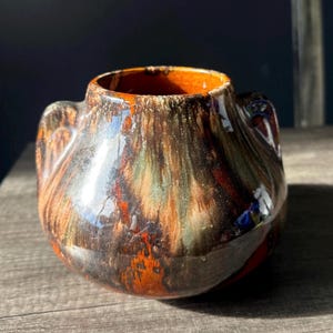 May include: A ceramic vase with a distinctive, rounded form. The vase is adorned with a multi-toned glaze in browns, oranges, and greens, giving a marbled appearance. It has two small handles and a wide opening, suitable for flowers or as a decorative item.