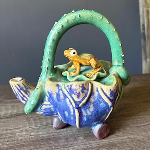 May include: A ceramic teapot with a frog figurine. The teapot has a blue and yellow body, a green handle and spout. A frog sits on a green leaf, adding a playful touch to this decorative piece. The teapot is a unique item.
