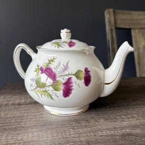 May include: A white porcelain teapot featuring a botanical illustration of purple thistle flowers with green leaves. The teapot has a rounded body, a matching lid with a small knob, a curved handle, and a spout, all accented with thin gold trim.