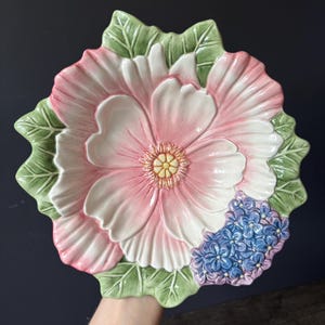 Może przedstawiać: Ceramiczny talerz w kształcie dużego różowego kwiatu z białymi brzegami płatków i żółtym środkiem. Talerz posiada zielone liście wokół krawędzi oraz skupisko małych niebieskich kwiatów w prawym dolnym rogu, całość z błyszczącym wykończeniem.