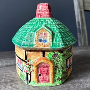 Peut inclure: Un pot en céramique en forme de cottage avec un couvercle amovible, doté d'un toit vert en bardeaux et d'une cheminée rouge. La base est beige avec des poutres noires, une porte rouge arquée, des fenêtres blanches et du lierre vert.