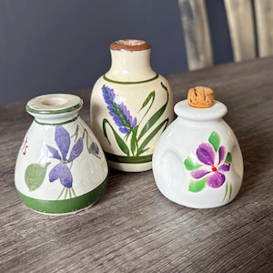 May include: Three hand-painted ceramic vases. The vases are white, with floral designs in purple and green. One vase has a cork stopper. The vases are on a wooden surface.