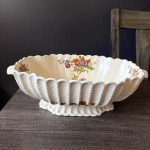 May include: Cream ceramic serving bowl with a scalloped rim and basketweave texture. The bowl has floral designs in purple, yellow, and red. It has a pedestal base and handles. The bowl is approximately 30cm in length.
