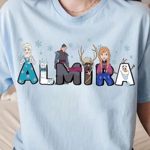 Puede incluir: Camiseta azul claro con un gr&aacute;fico de personajes de la pel&iacute;cula Frozen. Los personajes est&aacute;n dispuestos para deletrear el nombre "ALMIRA".
