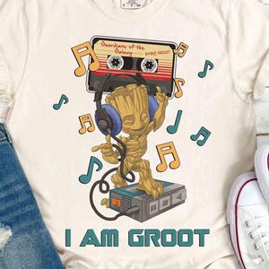 May include: Cream-coloured t-shirt featuring a cartoon Groot character wearing headphones and listening to music from a cassette tape. The text "I AM GROOT" is printed below the image, with musical notes scattered around.