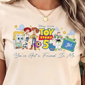 May include: Beige t-shirt featuring a colorful graphic of characters from the Disney Pixar movie Toy Story. The graphic includes the text "Toy Story 5" and "You've Got a Friend In Me".