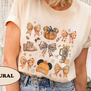 May include: A cream-colored t-shirt featuring a fall-themed graphic. The design includes various bows, pumpkins, boots, and a coffee cup, all in autumnal colors. The word "NATURAL" is printed on the bottom left of the shirt.