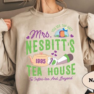 Peut inclure: Un sweat-shirt beige avec un motif graphique repr&eacute;sentant un personnage de dessin anim&eacute; portant un chapeau et tenant une th&eacute;i&egrave;re. Le texte sur le sweat-shirt est "Mrs. Nesbitt's 1995 Tea House To Infini-tea And Beyond". Le mot "NATURAL" est imprim&eacute; dans un rectangle en bas du sweat-shirt.