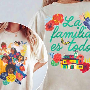 May include: Cream-colored t-shirt featuring a colorful illustration of several women surrounded by flowers and butterflies. The back of the shirt displays the text "La familia es todo" in green, along with a vibrant house graphic and floral accents.