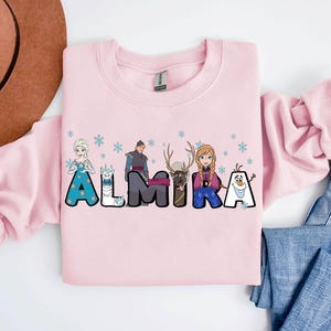 Puede incluir: Sudadera rosa con tem&aacute;tica de Frozen. Las letras del nombre ALMIRA est&aacute;n formadas por personajes de la pel&iacute;cula, incluyendo Elsa, Anna, Olaf, Kristoff y Sven.