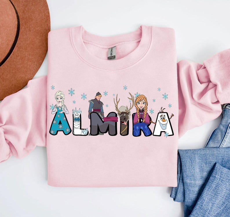 Puede incluir: Sudadera rosa con tem&aacute;tica de Frozen. Las letras del nombre ALMIRA est&aacute;n formadas por personajes de la pel&iacute;cula, incluyendo Elsa, Anna, Olaf, Kristoff y Sven.