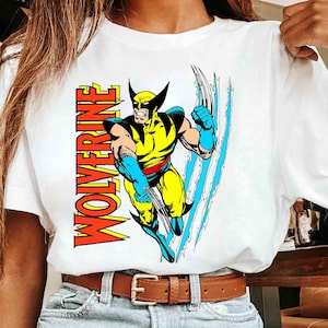 Op de afbeelding: Wit T-shirt met een kleurrijke afbeelding van Wolverine, een stripboekpersonage. Het ontwerp bevat het woord "WOLVERINE" in oranje en gele letters. Het personage wordt afgebeeld in een dynamische pose met blauwe klauwen.