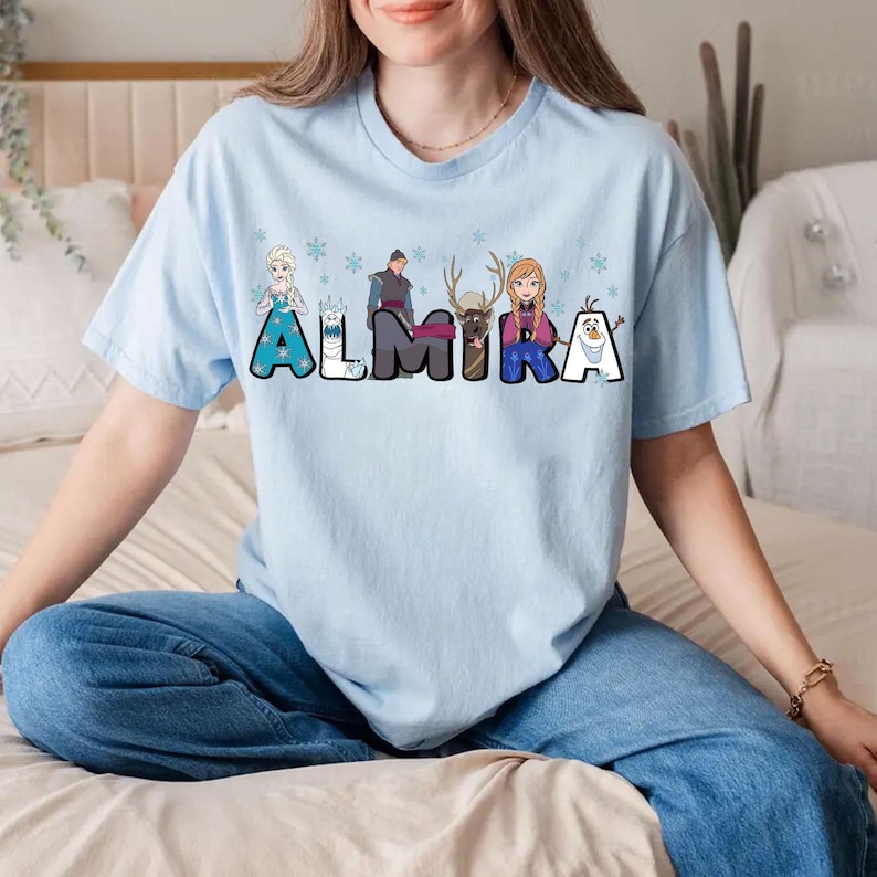 Puede incluir: Camiseta azul claro con un gr&aacute;fico de personajes de la pel&iacute;cula Frozen. Los personajes est&aacute;n dispuestos para deletrear el nombre "ALMIRA".