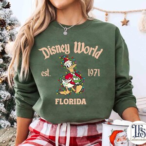 May include: Green crewneck sweatshirt with "Disney World" text and Donald Duck graphic wearing a Santa hat and Christmas lights. The text "est. 1971 FLORIDA" is also printed on the sweatshirt. The sweatshirt is a festive holiday design.