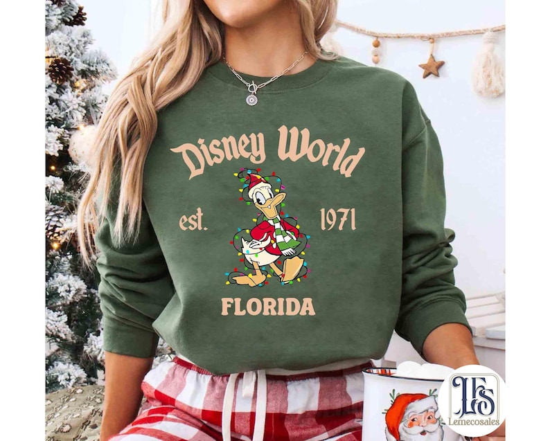 May include: Green crewneck sweatshirt with "Disney World" text and Donald Duck graphic wearing a Santa hat and Christmas lights. The text "est. 1971 FLORIDA" is also printed on the sweatshirt. The sweatshirt is a festive holiday design.