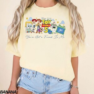 May include: A light yellow t-shirt featuring a graphic of characters from the Disney Pixar movie Toy Story 5. The design includes Woody, Buzz Lightyear, and other characters, with the text "You've Got a Friend In Me".