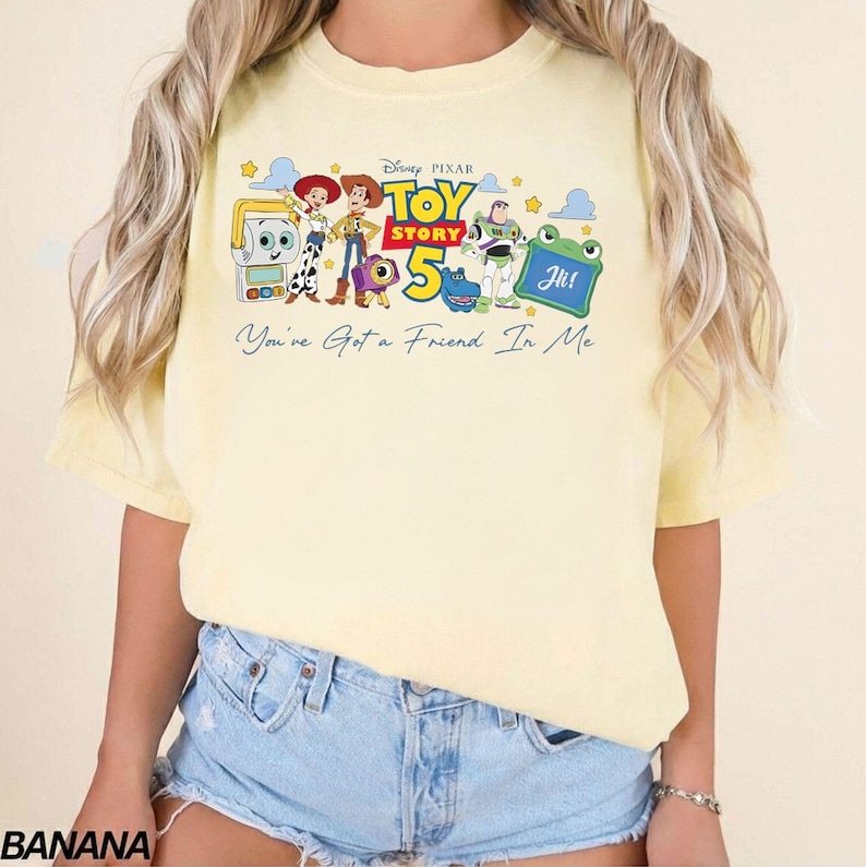 May include: A light yellow t-shirt featuring a graphic of characters from the Disney Pixar movie Toy Story 5. The design includes Woody, Buzz Lightyear, and other characters, with the text "You've Got a Friend In Me".