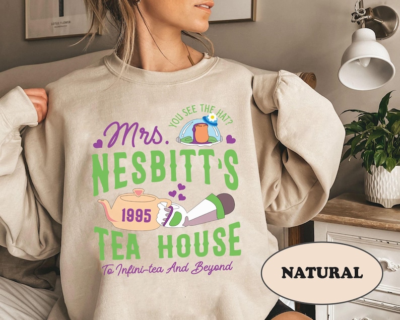 Peut inclure: Un sweat-shirt beige avec un motif graphique repr&eacute;sentant un personnage de dessin anim&eacute; portant un chapeau et tenant une th&eacute;i&egrave;re. Le texte sur le sweat-shirt est "Mrs. Nesbitt's 1995 Tea House To Infini-tea And Beyond". Le mot "NATURAL" est imprim&eacute; dans un rectangle en bas du sweat-shirt.